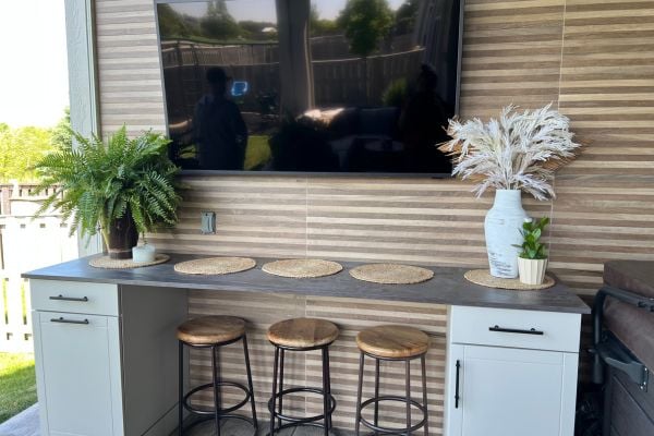 Wall mounted TV above a countertop with 3 barstools outside on the porch