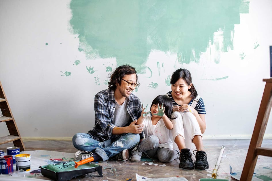 A family of 3 painting a wall in their home together. The young child has paint all over her hands while the parents smile