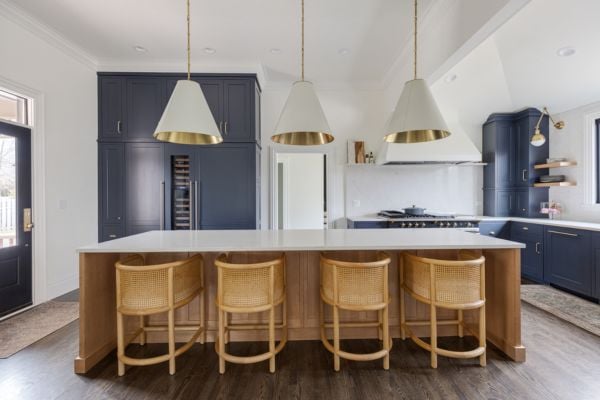 Kitchen center island in natural wood with white quartz countertops, featuring  cane barrel island bar seating, three large conical shade white and gold pendant lights and navy blue custom cabinetry