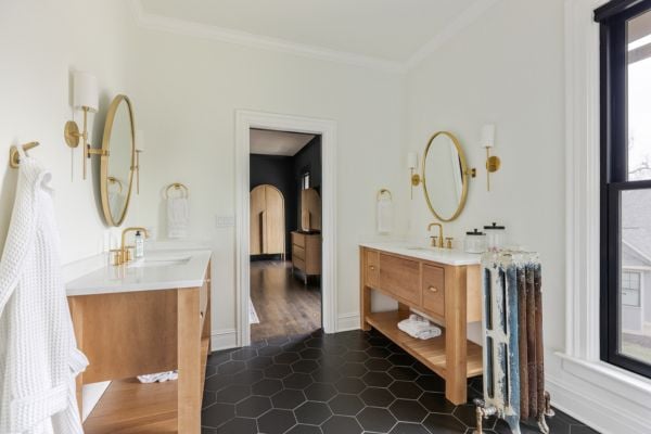 Primary Suite Bath with two single sink vanities in natural wood with white quartz countertops and gold accent and black hexagonal tile flooring