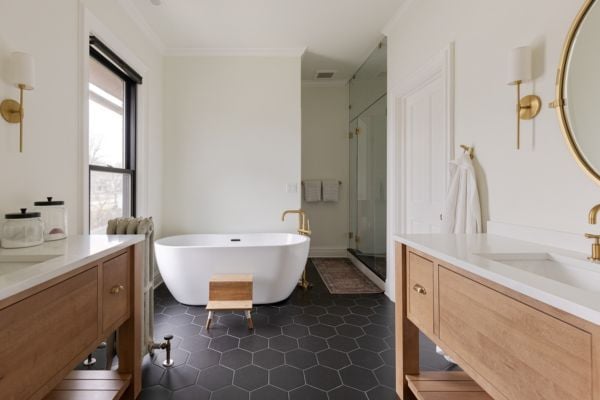 Primary Suite Bath with two single sink vanities in natural wood with white quartz countertops and gold accents, a large soaker tub, a steam shower and black hexagonal tile flooring