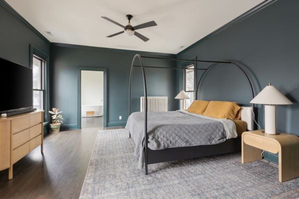 Primary Suite Bedroom, art deco inspired with metal arched bed and mid-century light wood dresser and night stand, featuring dark wood flooring, dark green walls and a globe light ceiling fan